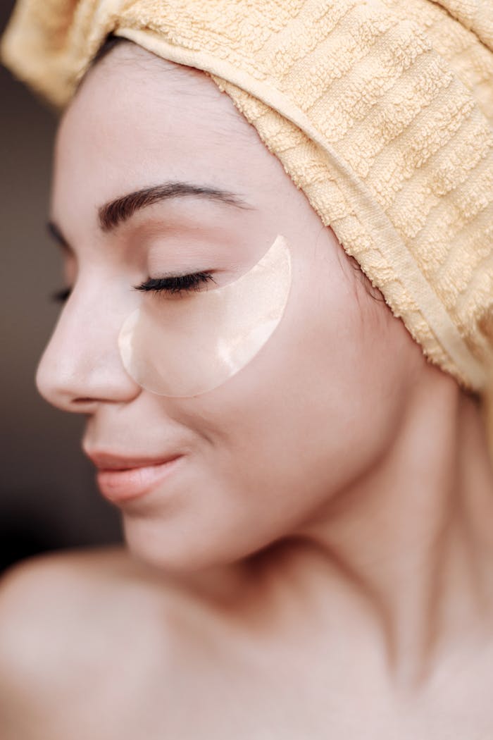 Profile of a woman with a towel on her head and under eye patch, emphasizing skincare.